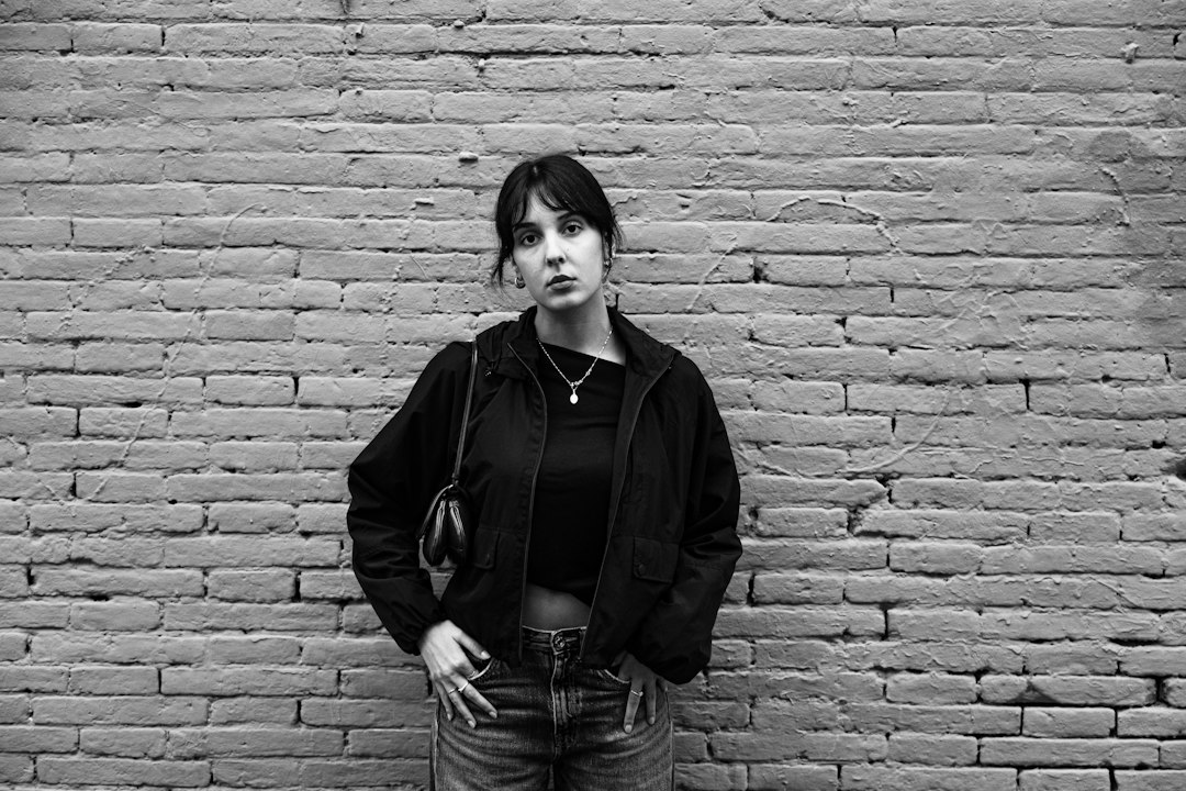 A woman in a black jacket and jeans standing against a textured brick wall